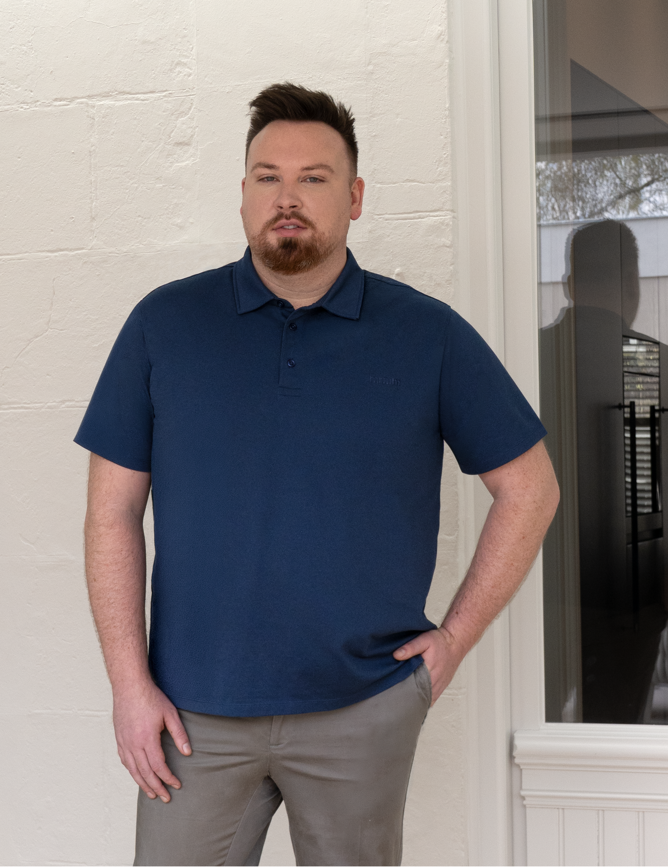 Man wearing a blue polo shirt and khaki pants standing in front of a white wall.