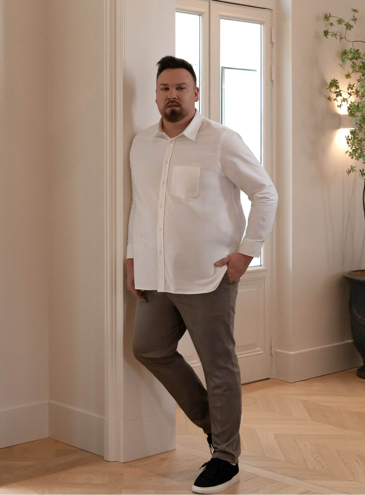Man wearing a white shirt and khaki pants standing in a room with a window and plant.